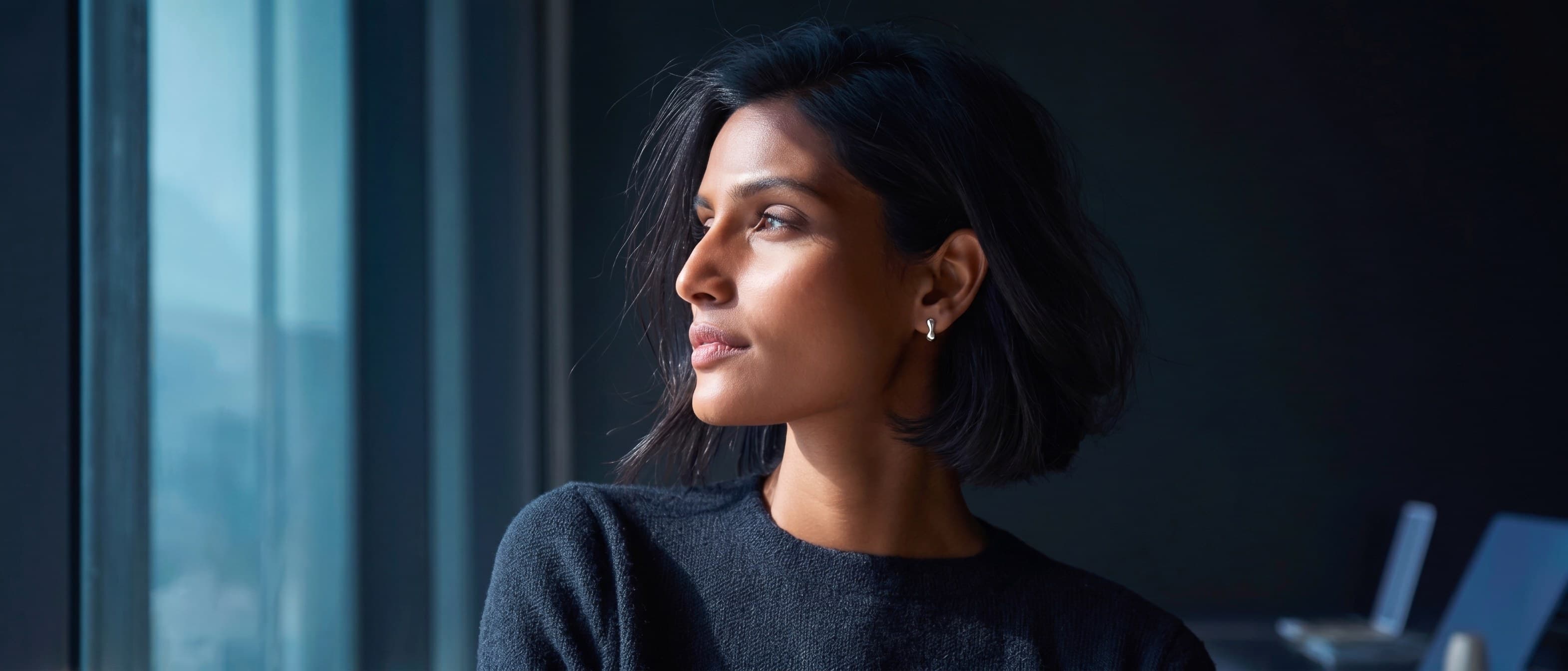 A woman at a desk by a window in a plain dark knit, turned to the light, one earring catching it.